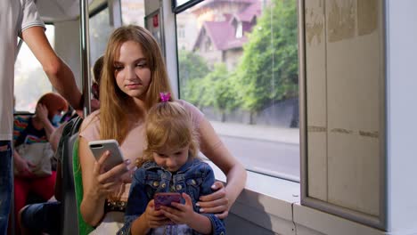 Child-girl-kid-with-mother-using-mobile-phone-chatting-in-social-media-on-crowded-tram,-bus-or-train