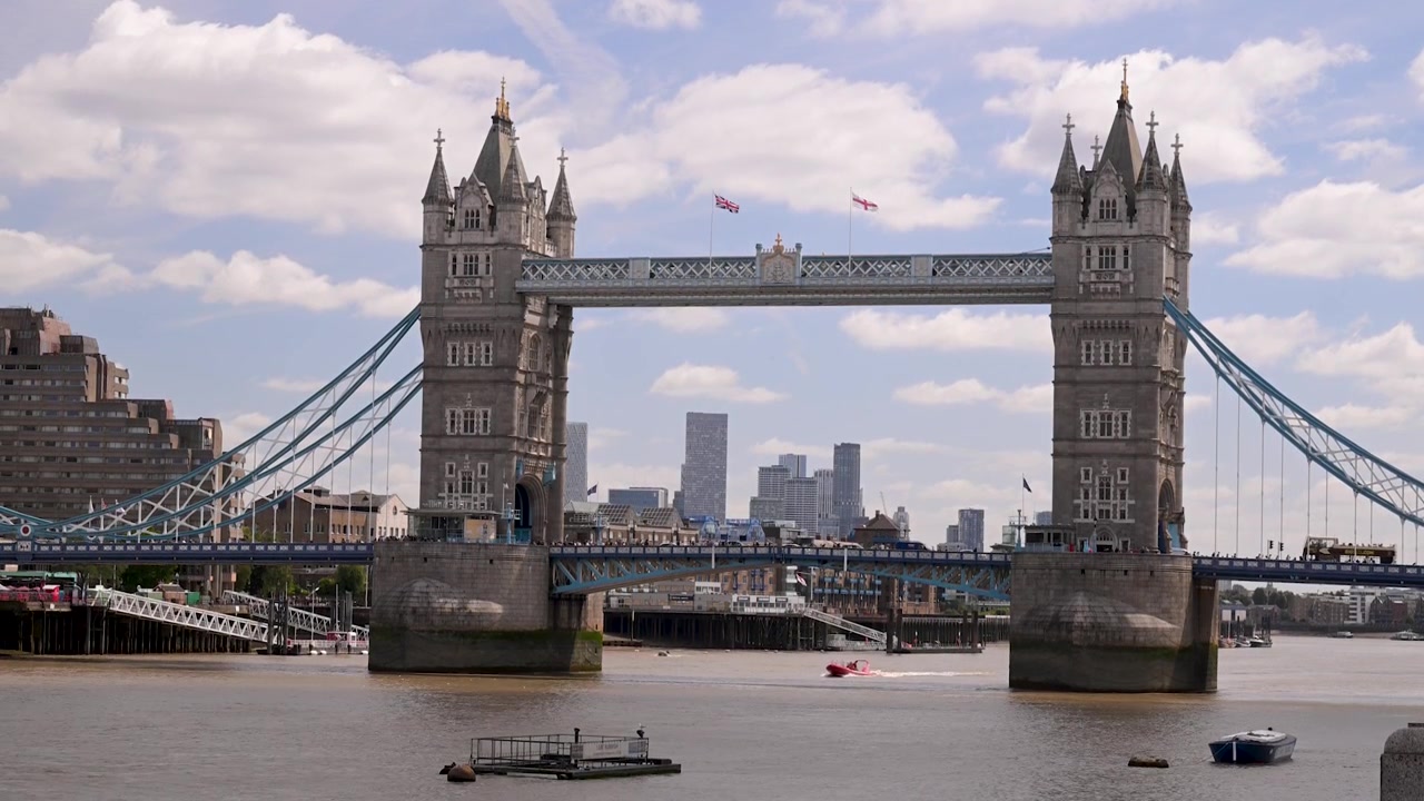 Tower Bridge strækker sig over Themsen på en solrig dag, mens små både bevæger sig på vandet med Londons skyline i baggrunden.