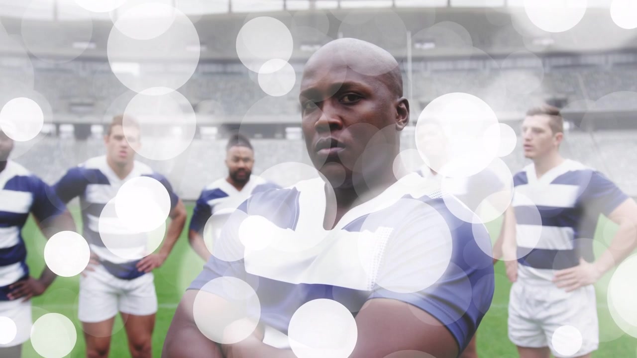 Male rugby player standing with arms folded on field, team, bokeh drifting obscuring for promo