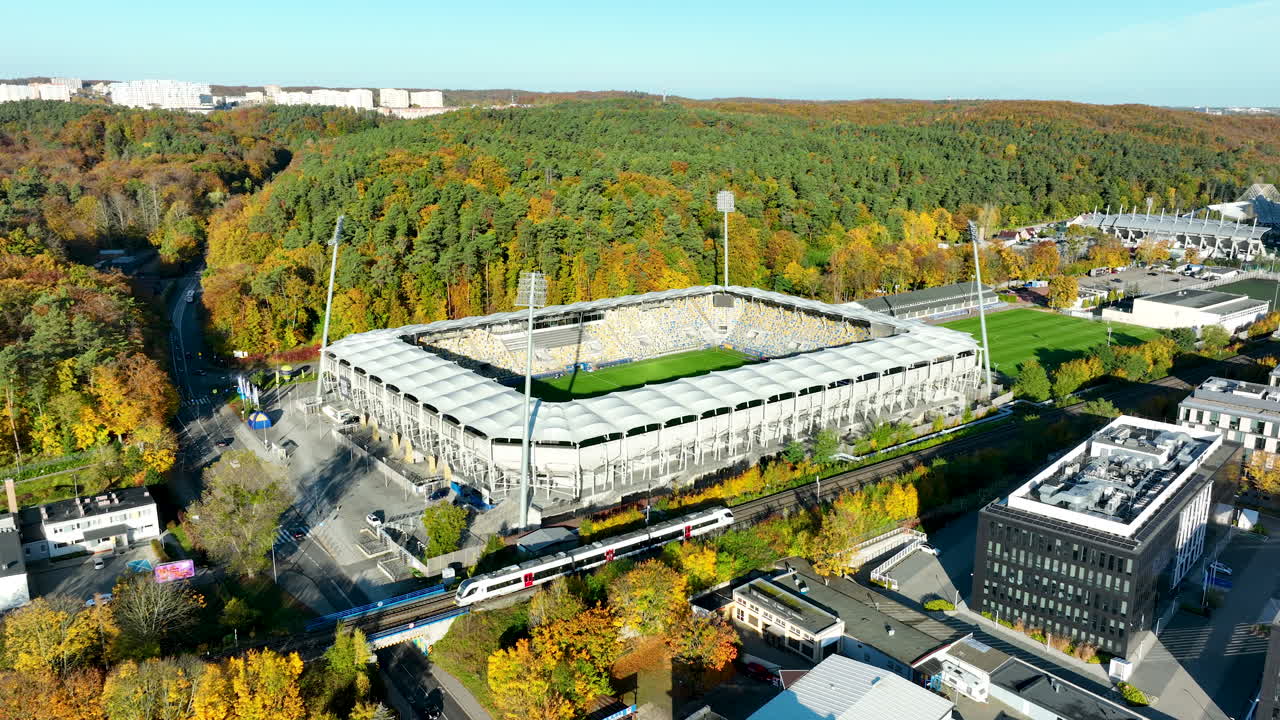 Drone footage of Gdynia city stadium surrounded by autumn trees and modern buildings in bright daylight