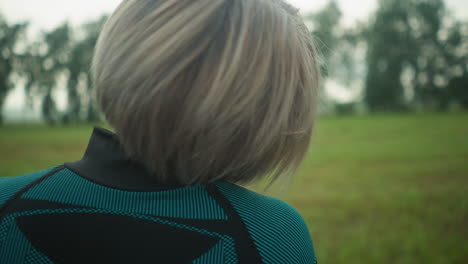 back view of woman in black and green suit, eyes closed, tilting her head slowly from right to left, stretching and relaxing her neck muscles outdoors in a peaceful grassy field under a cloudy sky