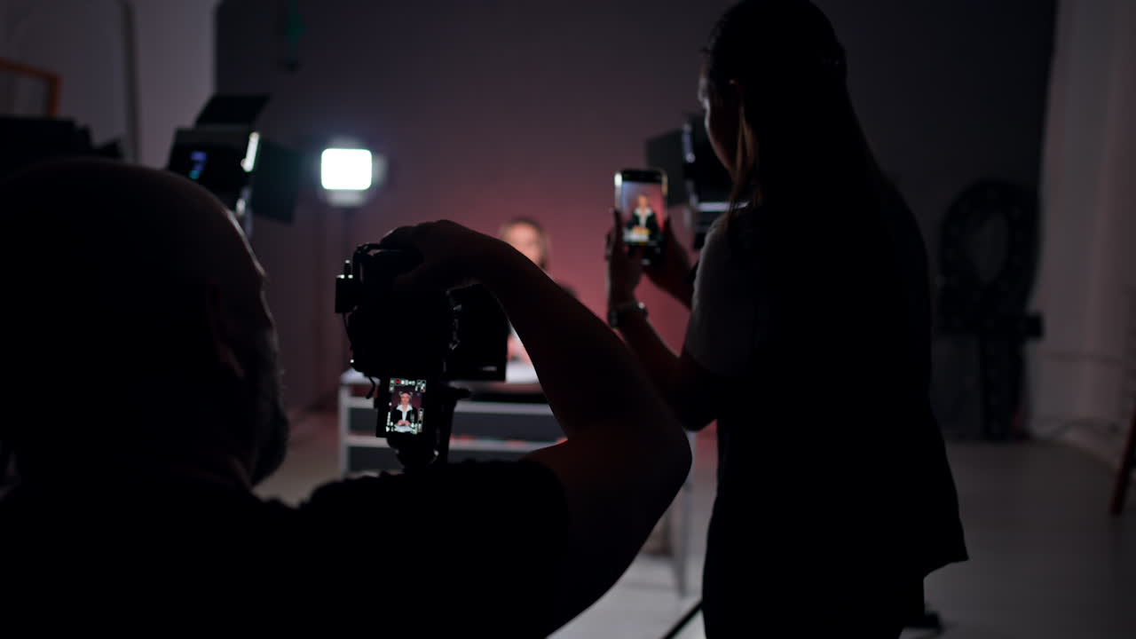 Blog crew works in the studio. Female blogger sits at desk. Man and woman filming a video on smartphone and professional camera.