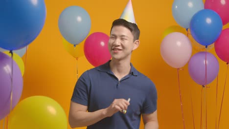 studio portrait of man wearing party hat celebrating birthday with balloons and party blower