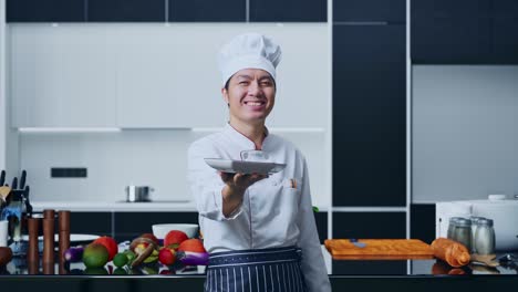 chef asiático sonriendo y dando un plato a la cámara mientras está de pie en la cocina de casa