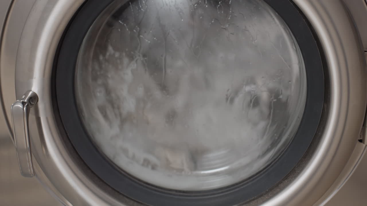 Zoom out transparent washer door reveals soapy water and fabric tumbling inside, droplets streaking glass, stainless rim in frame, continuous rotation during rinse cycle inside modern laundromat