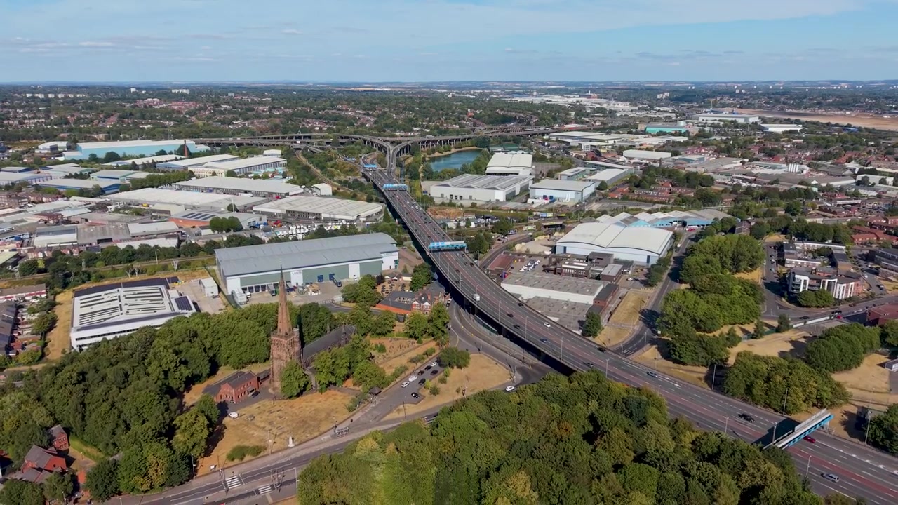Ripresa aerea della Spaghetti Junction di Birmingham che si allontana dallo svincolo M6 di Gravelly Hill, rivelando cavalcavia, flusso di traffico, magazzini, canali e l'espansione urbana sotto il cielo estivo.