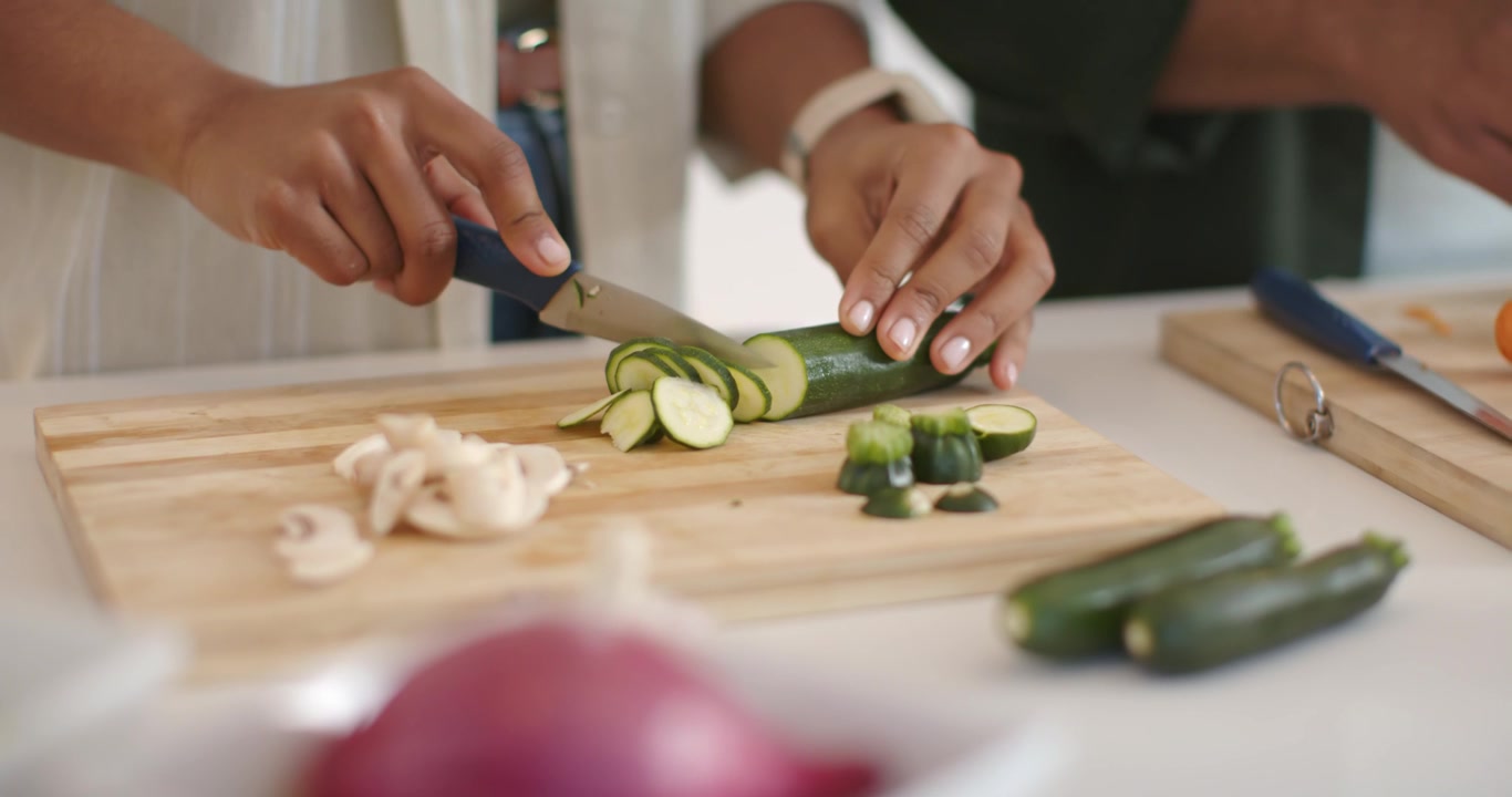 Coltello che scende mentre le mani tengono ferma la zucchina e la affettano a rondelle su un tagliere di legno, preparando il pasto