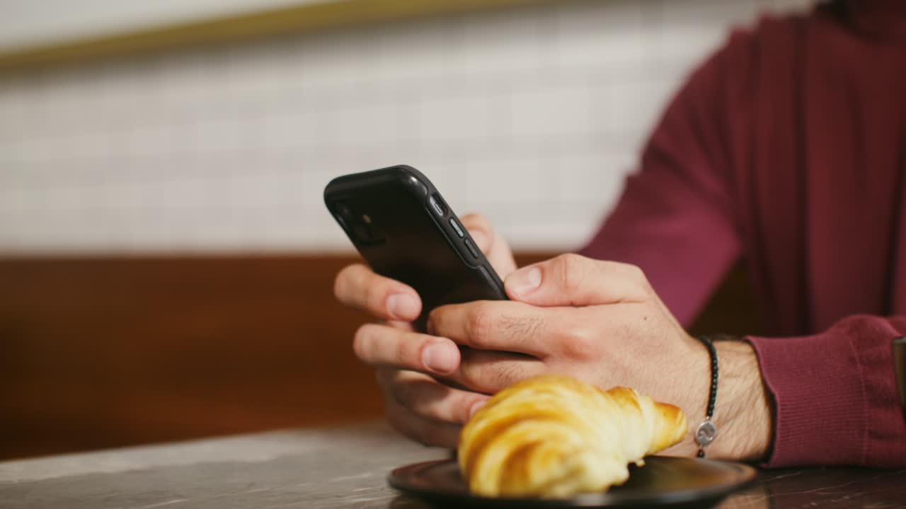 un hombre disfrutando de un café y un croissant mientras usa su teléfono inteligente en una cafetería.