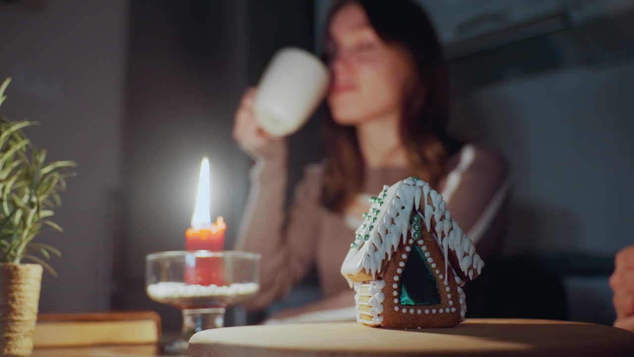 색 아이싱으로 세부적인 진저브레드 집 에 불타는 은 불의 클로즈업. 흐릿한 배경에서 여성은 에 머리를 고 차를 마시고 있습니다.