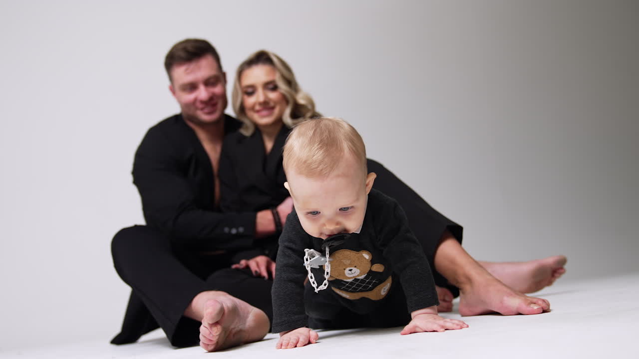 Cute blond baby with pacifier in mouth crawl by the floor. Happy parents sitting behind look at their child. Low angle view. White backdrop.