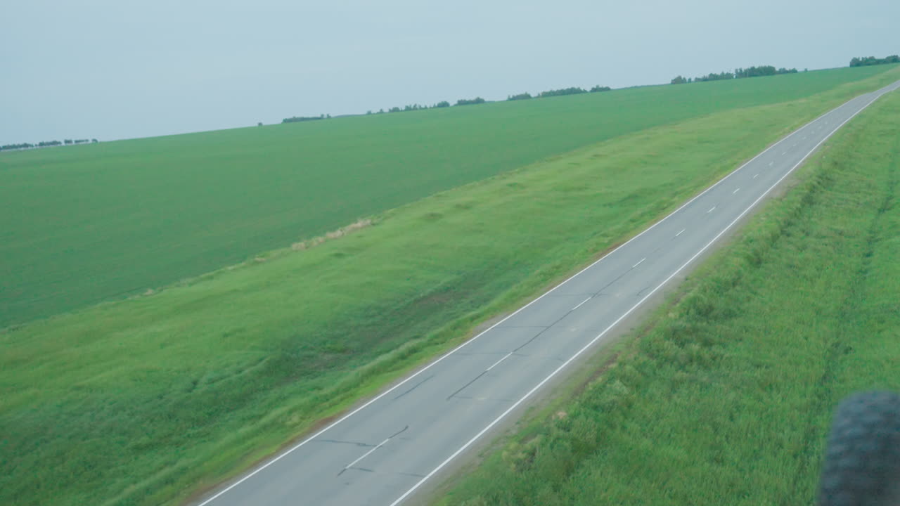 overhead drone capturing straight rural road cutting through lush green farm land under soft daylight white dashed markings guiding visual corridor toward distant horizon in serene aerial perspective