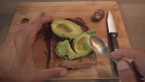 person smashing fresh avocado on toasted bread by teaspoon