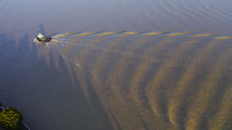 Aerial-birds-eye-view-tugboat-passing-by-a-commercial-river-causing-waves-on-a-twilight-sunset-muddy-water-just-returning-to-the-port-dock-to-haul-another-boat-into-the-sea-for-import-export-contracts