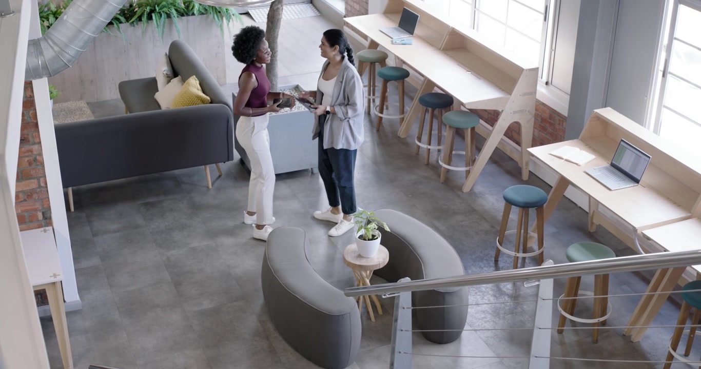 Diverse female coworkers descending mezzanine, exchanging device, reviewing contents while walking