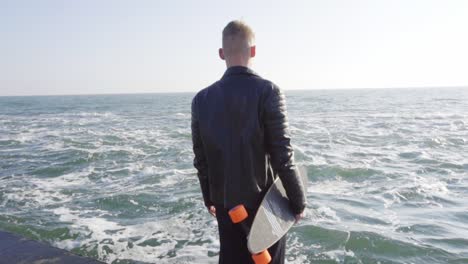 young man in black leather jacket holds his longboard by the sea