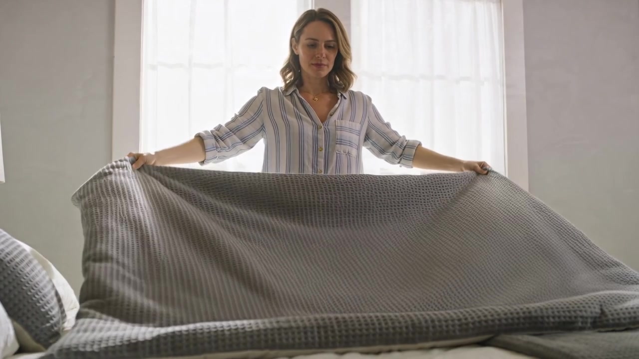 Scuotendo, una donna in camicia a righe stende, tira e liscia la coperta ai piedi del letto per sistemarla.