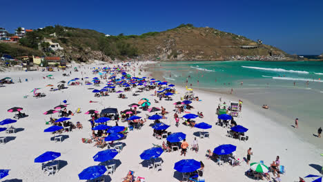 drone captures vibrant beach umbrellas, white sand, swimmers in blue waters, and scenic mountains in the backdrop in arraial do cabo