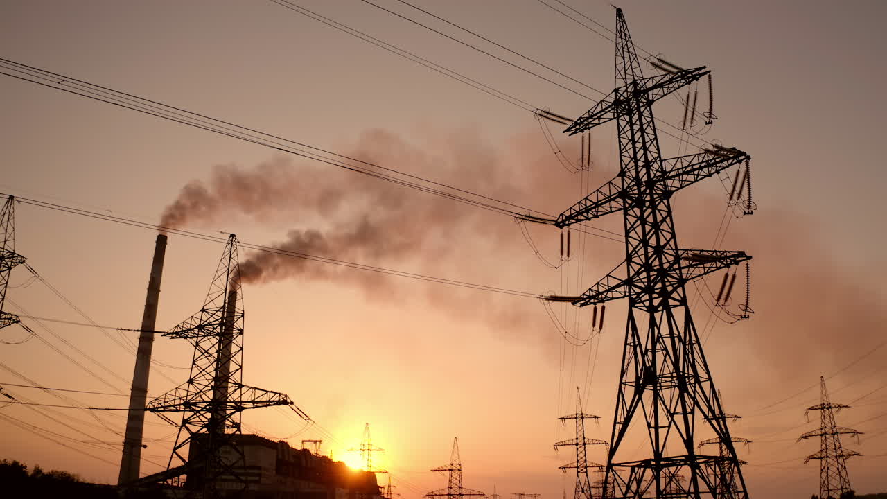 Electricity pylons against smoke stacks at sunset. High-voltage electric towers on the evening sky. Factory pipes with smoke pollute the air. Motion camera down.