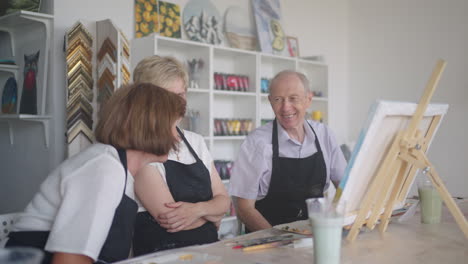 high angle view of cheerful senior friends painting on canvas. senior woman smiling while drawing with the group. seniors attending painting class together. senior men having fun painting in art class