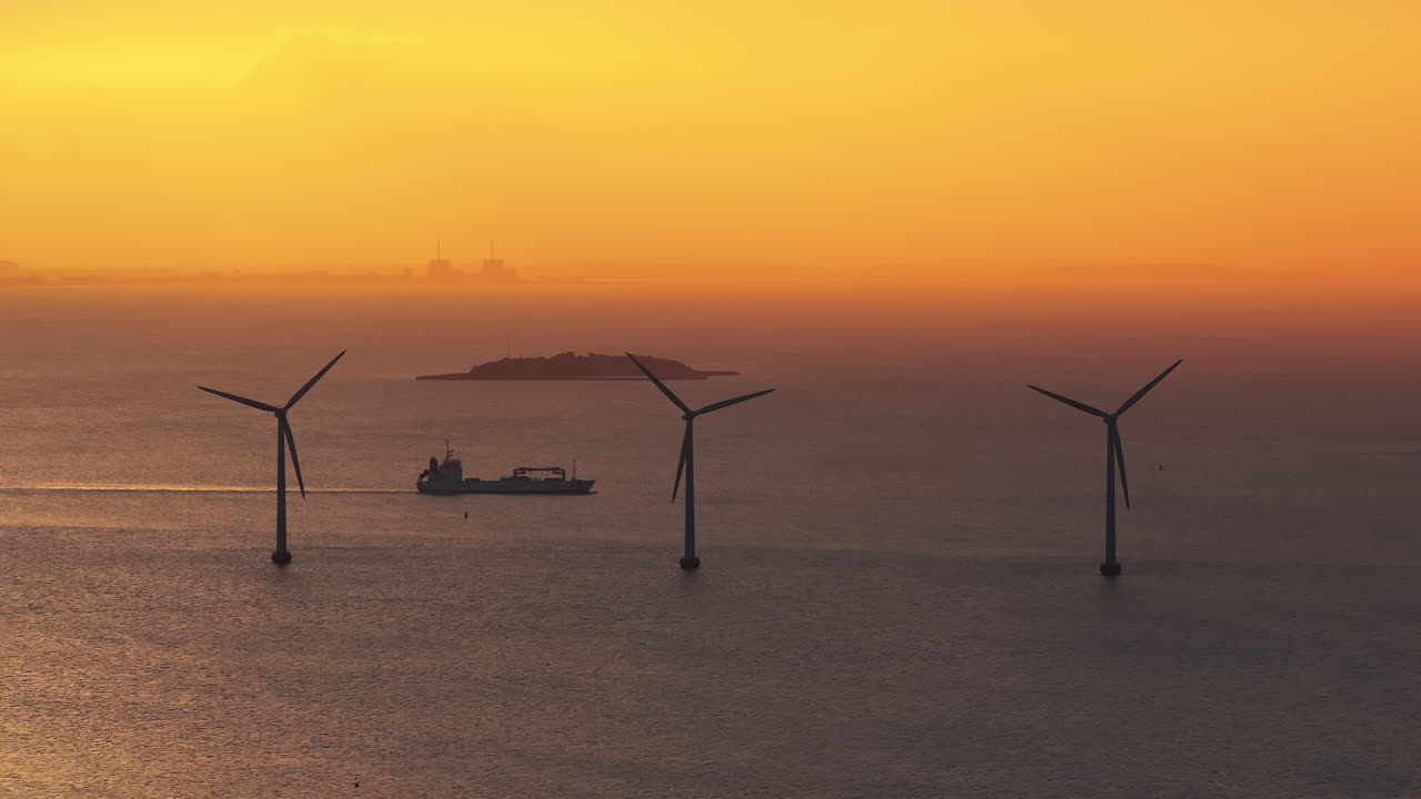 Aerial drone view of cargo ship sailing past offshore wind turbines in the Oresund Strait during a golden sunset in Copenhagen, Denmark