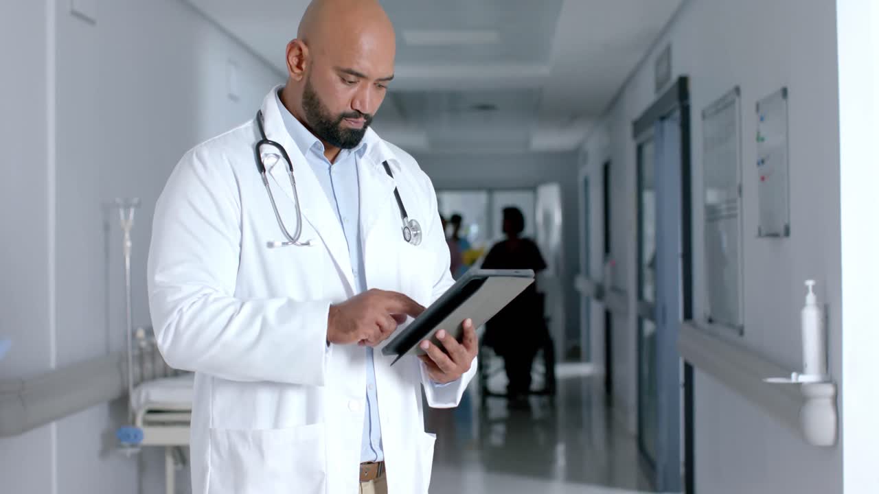 Biracial male doctor wearing lab coat and stethoscope, using tablet in corridor, slow motion