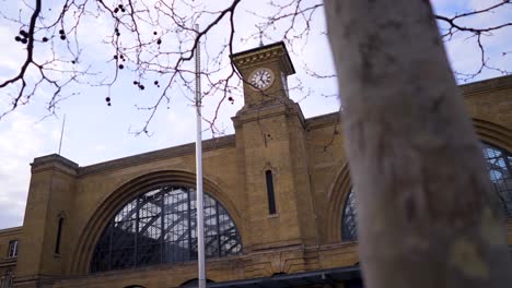 outside of king's cross train station in london, clock ticking, with a tree trunk in the foreground, on a sunny day with light clouds