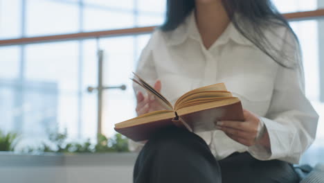 close up of young woman seated indoors flipping pages of open book with tattooed hand, wearing white shirt, surrounded by natural lighting and blurred modern mall interior in soft background