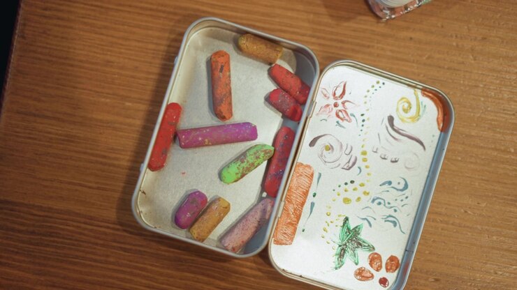 Close up of colorful pastels in metal tin with hand drawn designs on lid resting steadily. scene features vibrant chalk pieces and artistic doodles inside open container set against wooden