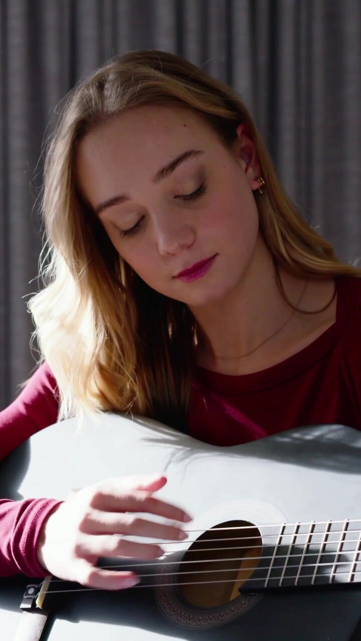 Vertical video: Strumming woman in burgundy top playing dark acoustic guitar on bed