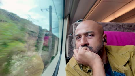 south asian man with earphones gazing out a train window, reflecting on his journey. scenic landscape visible through the glass