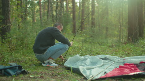 young man squats in forest attempting to insert flexible tent pole through fabric ring while setting up camping tent, sunlight filters through trees, casting warm glow over natural outdoor scene