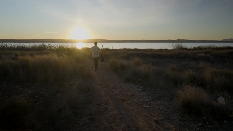 man walking with guitar at sunset by the lake