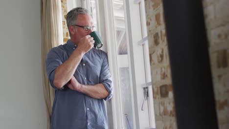 Happy-caucasian-man-wearing-glasses,-looking-through-window-and-drinking-coffee