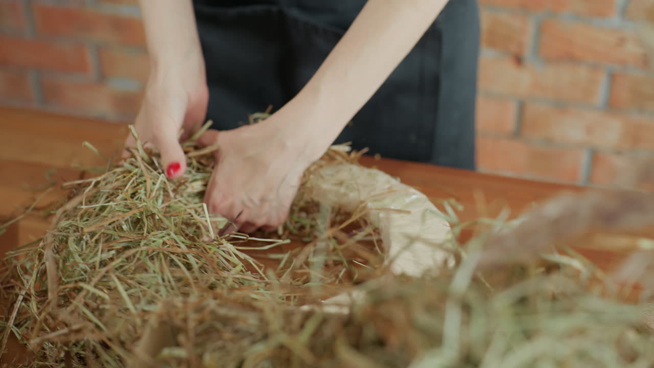 Florist arranges dried grass and flowers on circular base, focusing on detail of floral craft process with hydrangea blossoms and natural textures, emphasizing handmade rustic decoration in workspace