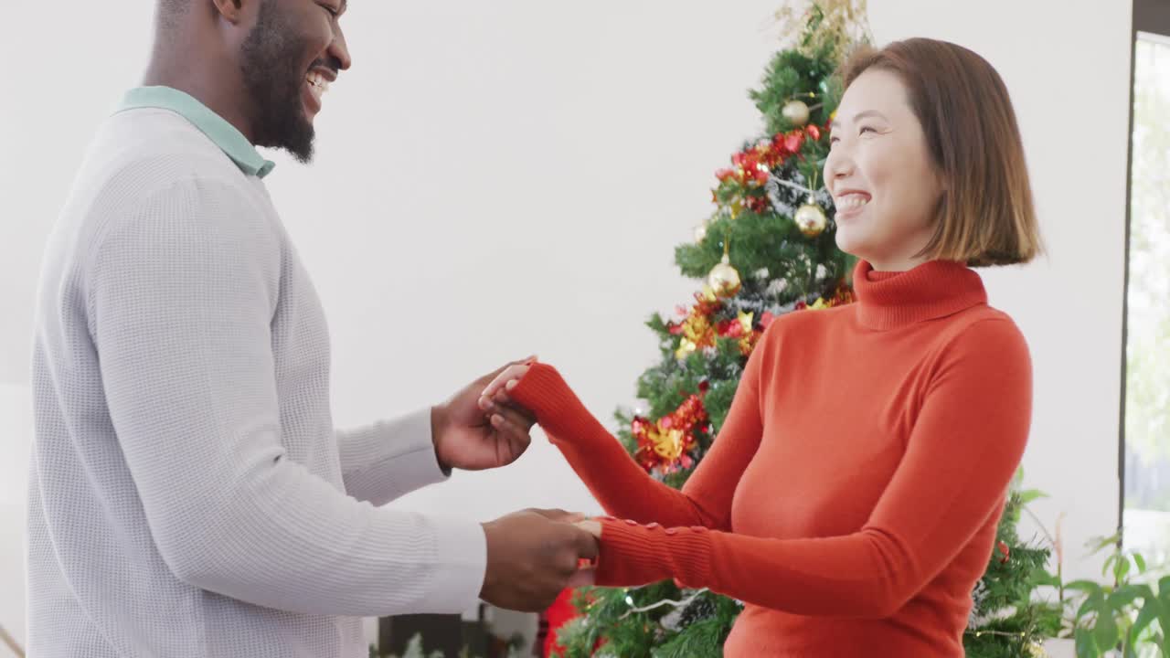 Video of happy diverse couple dancing beside christmas tree at home