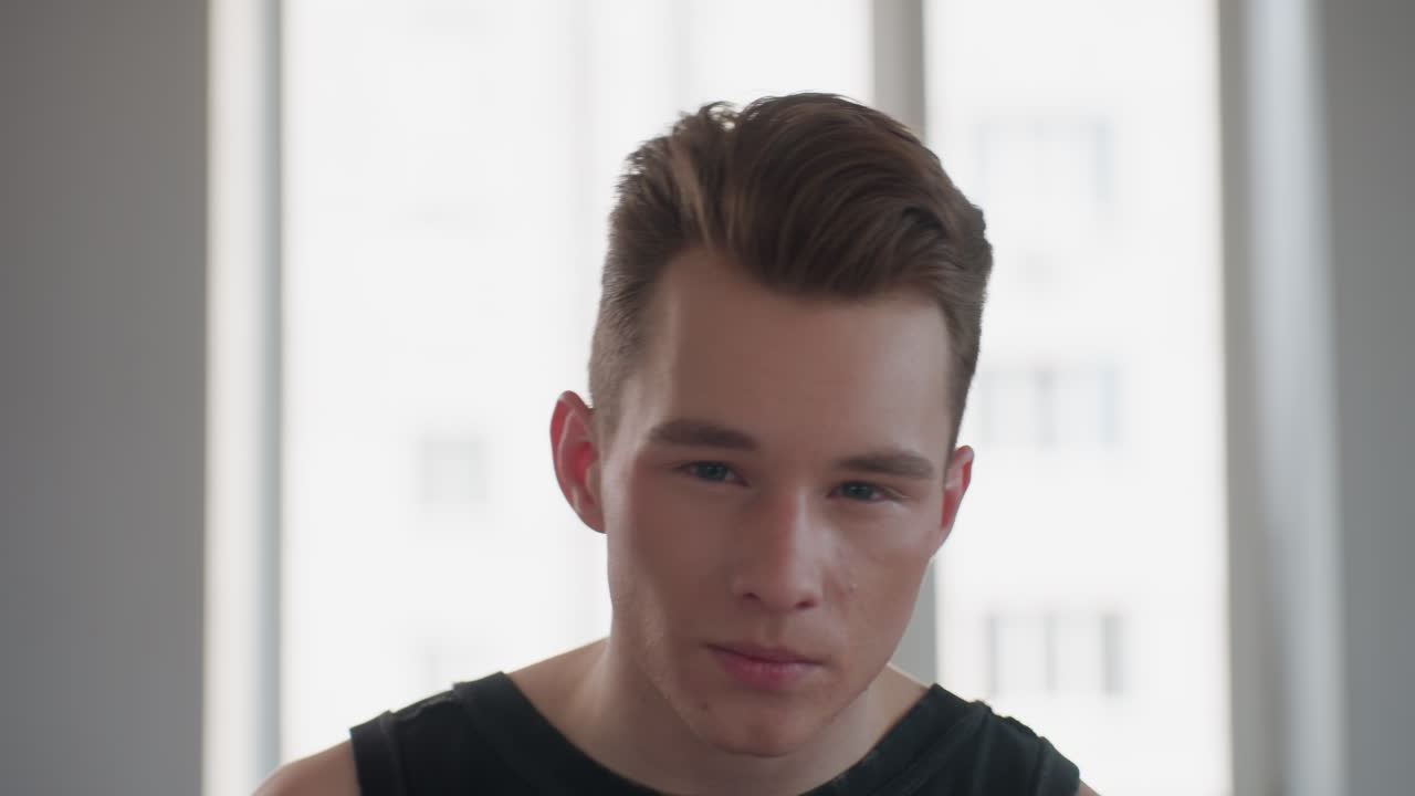 handsome tattooed man in sleeveless signet sits before bright window mirror, holds black makeup sponge, delicately applies powder to cheeks with poised expression