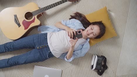 top view of asian teen girl enjoys playing smartphone while lying on carpet on the floor at home