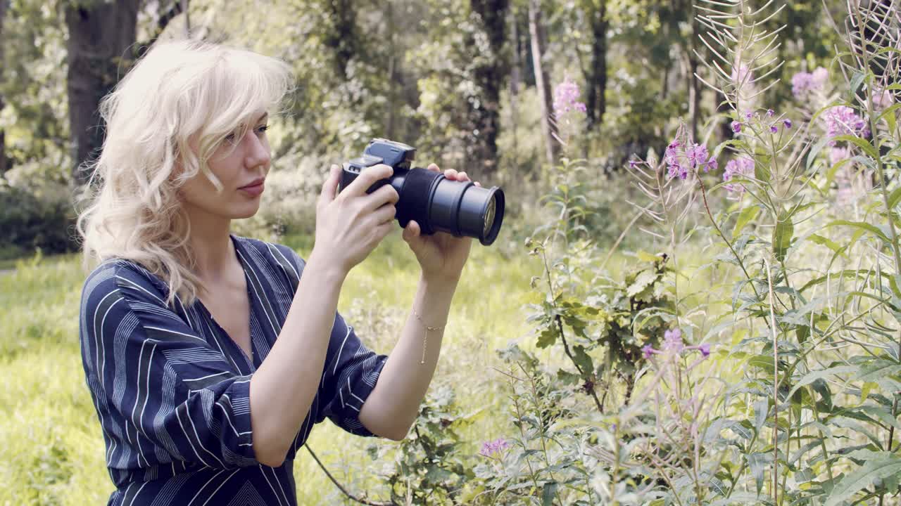 mujer rubia usa una cámara grande y una lente para tomar fotografías de flores silvestres en un parque