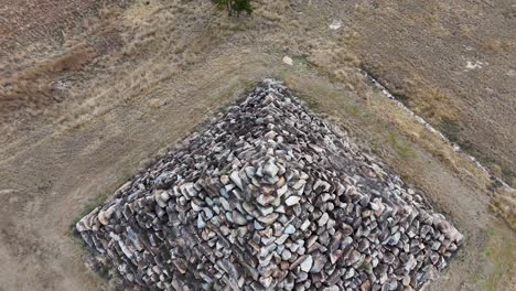a drone camera ascends above a large stone pyramid structure in a dry, grassy field under natural daylight, revealing geometric patterns and rural surroundings