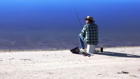 pescador se sienta en un balde boca abajo en una playa y arroja su línea de pesca