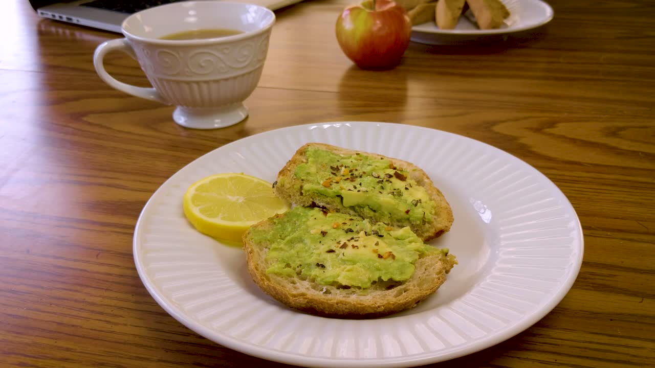 Close up of a plate of avocado toast on a wooden table with cup of tea, laptop, and apple in the background