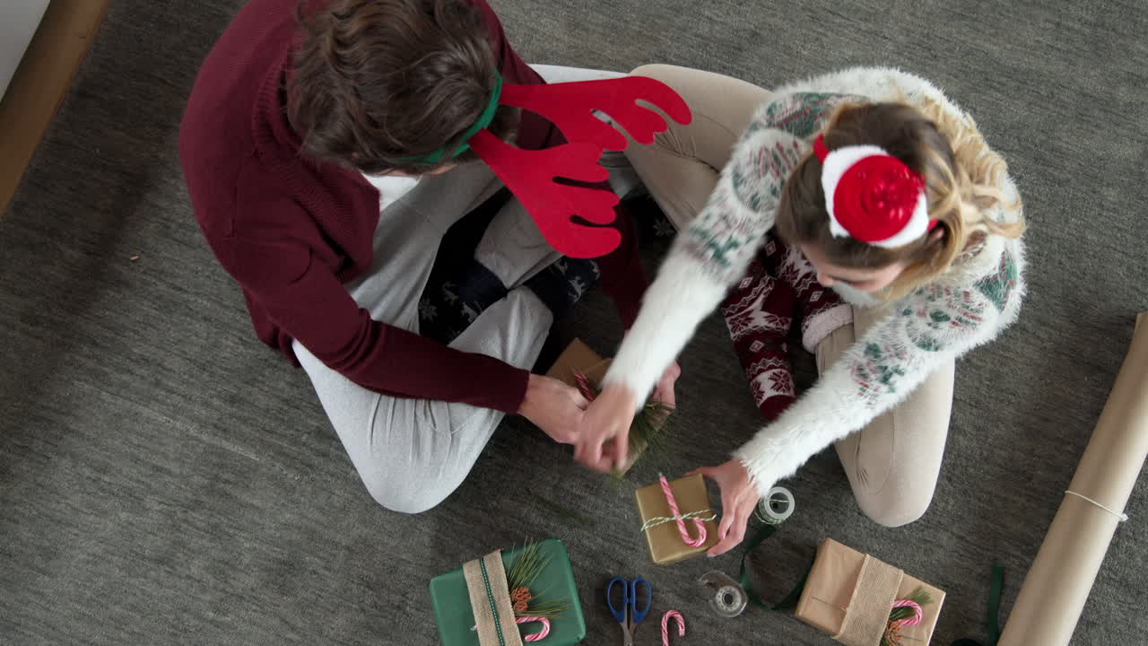 Couple wrapping Christmas gifts together on floor, wearing festive holiday attire