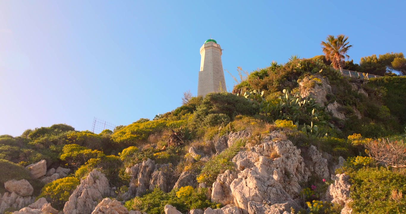Inquadratura dal basso che si inclina verso un faro bianco in cima a una collina rocciosa mediterranea con fiori selvatici gialli e palme.