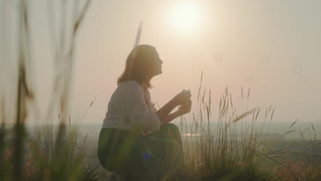 silhouetted woman crouches in tall grass blowing bubbles toward glowing sky as sun sets, with gentle light casting warm tones across landscape and bubbles floating in air