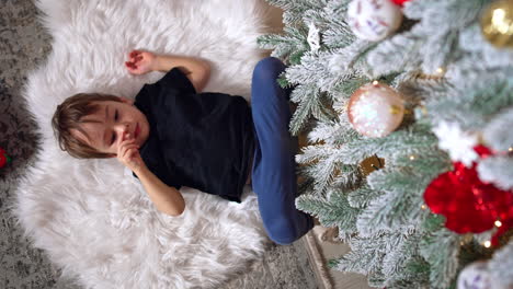 two year old toddler lies on white soft fluffy plaid on the floor. baby boy looks at christmas tree. vertical screen.