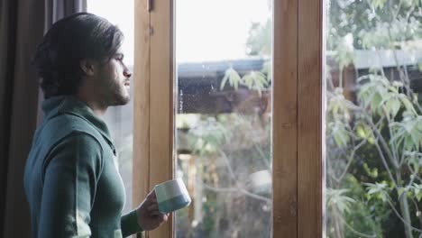 thoughtful caucasian man with cup of coffee and looking out window at home, copy space, slow motion