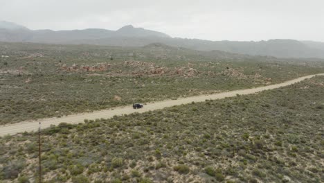 el dron vuela alto cerca de una carretera y sigue un vehículo fuera de carretera negro a vista de pájaro - paisaje desértico en el área silvestre de cederberg en sudáfrica - la cordillera se puede ver en el horizonte