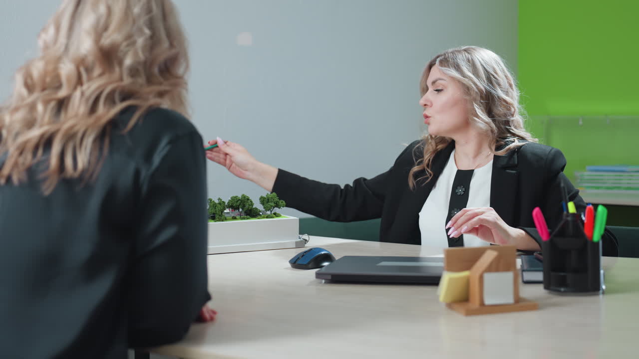 Realtor presenting residential development project to client using pencil to highlight features of architectural model during professional discussion, both engaged in office setting