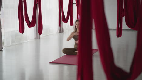 meditation instructor seated cross legged on red mat places hand on chest and other on stomach while closing eyes in calm breathing exercise amid hanging silk hammocks in bright yoga studio