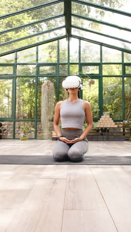 vertical video: practicing yoga, woman wearing vr headset and sitting on yoga mat in glass house